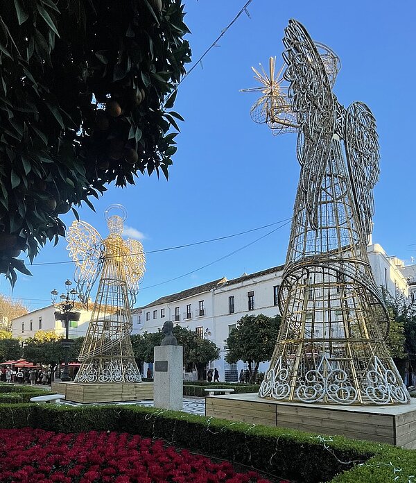 Weihnachtlich geschmückter Platz mit zwei großen beleuchteten Engelsfiguren aus Metall, gepflegten Buchshecken und einem Beet voller roter Blumen vor hellen historischen Häusern unter blauem Himmel.