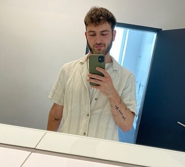 Ein junger Mann mit Bart steht mit einem grauem Smartphone vor einem Spiegel und fotografiert sich.