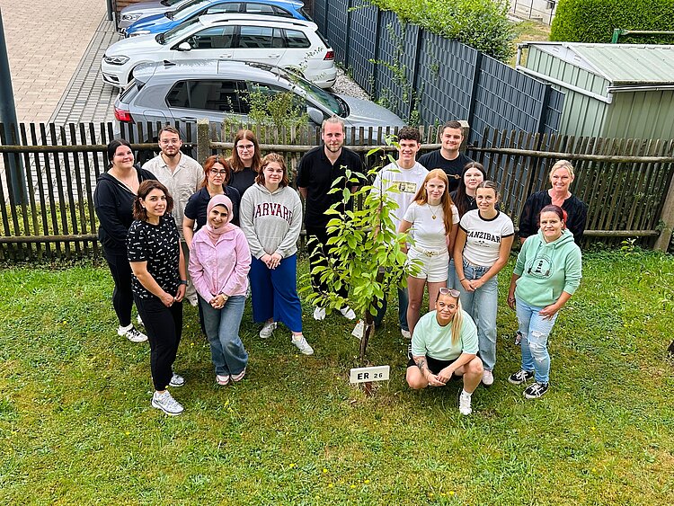 Gruppe von Jugendlichen und Erwachsenen steht auf einer Rasenfläche rund um einen neu gepflanzten jungen Baum und blickt gemeinsam in die Kamera.