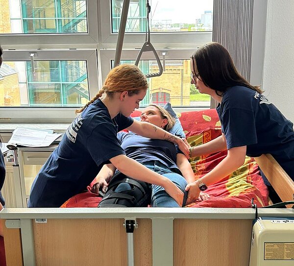 Zwei Personen in blauen T-Shirts helfen einer liegendem Patientin in einem Krankenhausbett. Die Patientin trägt ein blaues T-Shirt und hat eine Beinschiene an. Im Hintergrund sind große Fenster mit Blick auf Gebäude und Bäume sichtbar.
