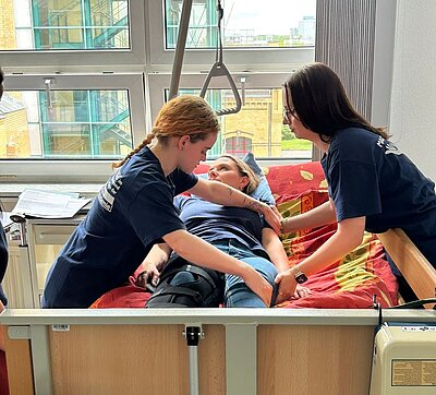Zwei Personen in blauen T-Shirts helfen einer liegendem Patientin in einem Krankenhausbett. Die Patientin trägt ein blaues T-Shirt und hat eine Beinschiene an. Im Hintergrund sind große Fenster mit Blick auf Gebäude und Bäume sichtbar.