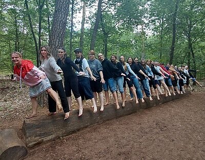 Eine große Gruppe von Menschen balanciert auf einem Bein mit nackten Füßen auf einem liegenden Baumstamm in einem Wald.