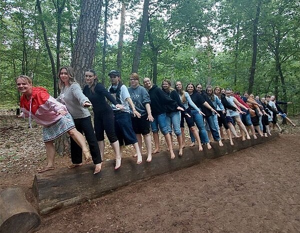 Eine große Gruppe von Menschen balanciert auf einem Bein mit nackten Füßen auf einem liegenden Baumstamm in einem Wald.