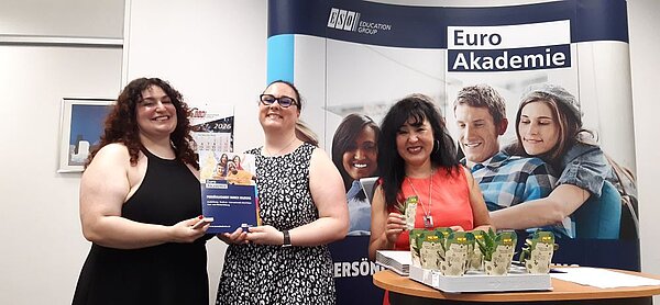 Drei lächelnde Frauen stehen vor einem Roll-up der Euro Akademie. Zwei der Frauen halten zusammen eine Informationsbroschüre in der Hand, während auf dem Tisch vor ihnen kleine Töpfe mit grünen Pflanzen und weitere Informationsmaterialien zu sehen sind.