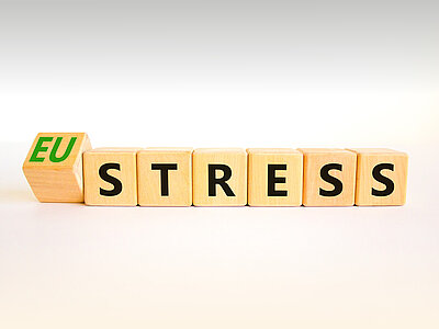 Holzblöcke mit Buchstaben, die das Wort 'EU STRESS' bilden. Der Block mit den Buchstaben 'EU' ist grün und steht leicht versetzt von den anderen Blöcken, die das Wort 'STRESS' in schwarzen Buchstaben zeigen. Die Blöcke sind auf einem hellen Hintergrund platziert.