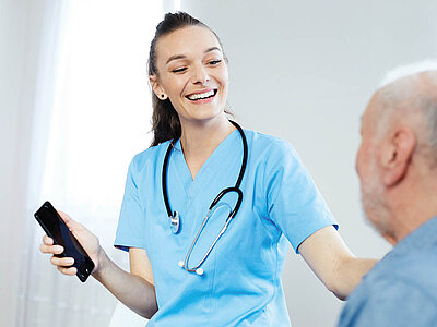 Auf dem Bild sieht man eine Krankenpflegehelferin, die einem Patienten etwas auf dem Handy zeigt. Die Interaktion findet in einer medizinischen Umgebung statt.