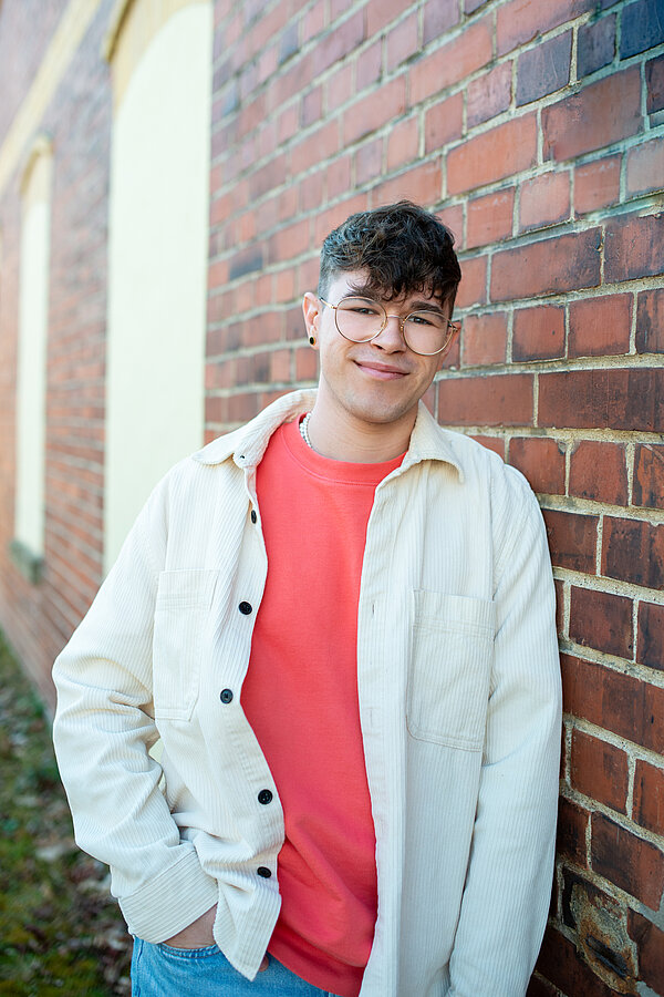 Ein junger Mann lehnt seitlich an einer Backsteinmauer und lächelt in die Kamera.