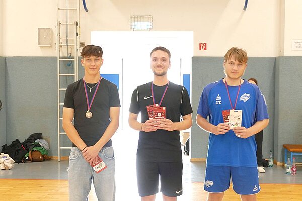 Drei junge Männer stehen nebeneinander in einer hellen Turnhalle und tragen Medaillen um den Hals, während sie Karten in den Händen halten und in die Kamera lächeln. Der Mann in der Mitte trägt ein schwarzes Sportoutfit, während die anderen beiden Sportkleidung in Blau oder Schwarz tragen.