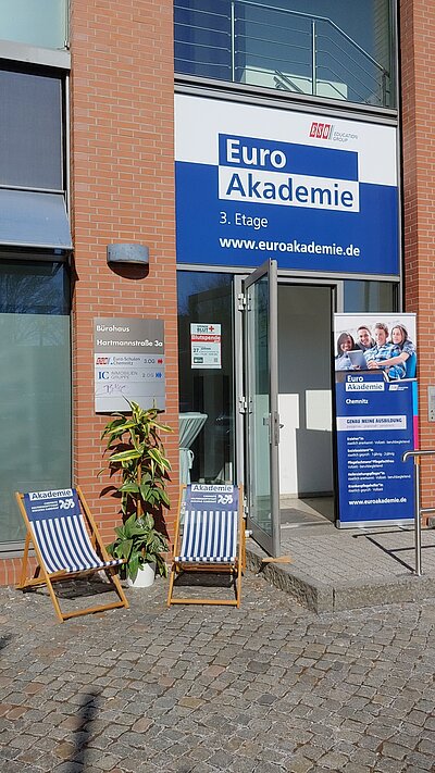 Der Eingangsbereich der Euro Akademie in der Hartmannstraße (3. Etage) befindet sich in hellem Sonnenschein. Zwei Strandstühle im blau-weißen Euro Akademie-Design laden zum Verweilen ein. Ein Roll-up neben der Tür klärt über die Angebote der Schule auf.