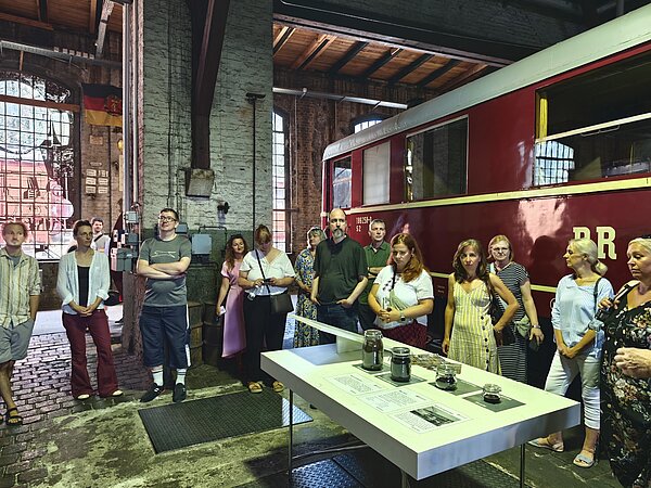 Eine Gruppe von Menschen steht in einer Halle mit Backsteinwänden und großen Fenstern. Vor ihnen befindet sich ein langer Tisch mit Ausstellungsstücken, darunter Gläser und Informationen. Im Hintergrund ist ein roter Zug mit der Aufschrift 'DR' zu sehen. Die Atmosphäre wirkt wie eine Führung oder Ausstellung.