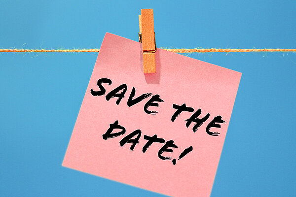 Die Aufschrift "Save the Date!" steht wie handschriftlich geschrieben auf einem rosafarbenen Notizzettel, der mit einer Holzwäscheklammer an eine Kordel gehängt ist. Der Hintergrund ist blau.