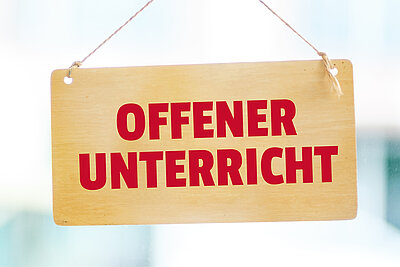 Ein hölzernes Schild mit der Aufschrift ‚OFFENER UNTERRICHT‘ in roten Großbuchstaben hängt an einer Schnur.