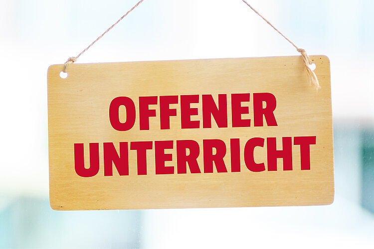 Ein hölzernes Schild mit der Aufschrift ‚OFFENER UNTERRICHT‘ in roten Großbuchstaben hängt an einer Schnur.