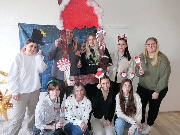 Eine Gruppe von neun Studierenden posiert vor einer weihnachtlich dekorierten Fotowand, einige halten Weihnachtsrequisiten wie Hüte, Rentiere und Santa-Figuren in der Hand.