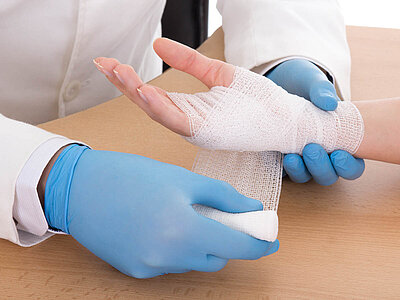 Ein Arzt in blauen Handschuhen verbindet eine Hand mit einem weißen Verband. Die Patientin ist entspannt, während der Arzt sorgfältig vorgeht.