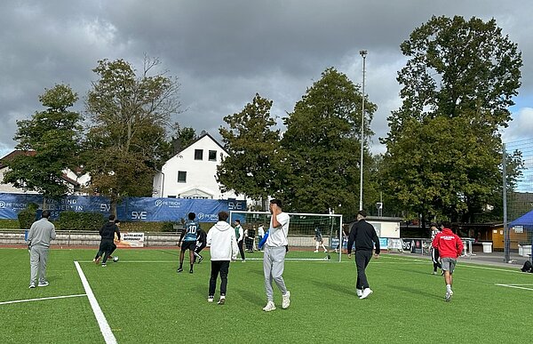 Together in motion – Bewegung, Spaß und Teamgeist Eine Gruppe von Menschen spielt auf einem Kunstrasenplatz Fußball. Einige Spieler stehen oder laufen auf dem Platz, während im Hintergrund ein Tor und Bäume zu sehen sind.