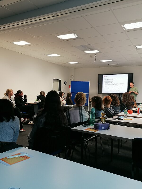 Seminarraum mit etwa 20 Teilnehmenden; Dozent*in erklärt Inhalte vor projizierter Folie.