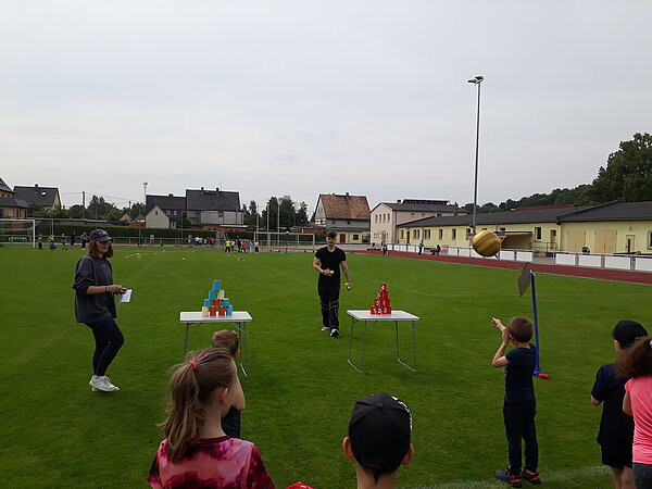 Unser Motto zum Sportfest: "Auf zu neuen Galaxien!" Ein Wurfspiel mit bunten Bechern auf einem Tisch wird von zwei jungen Erwachsenen betreut, während Kinder aufmerksam zuschauen.