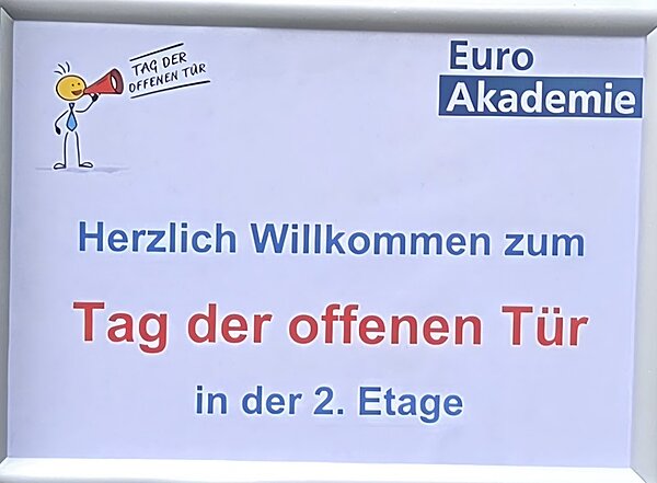 Ein weißen Schild, mit Euro Akademie Logo in der oberen rechten Ecke, mit einem Cartoon-Mann mit Megaphon und dem Text Herzlich willkommen zum Tag der offenen Tür in der 2. Etage. 