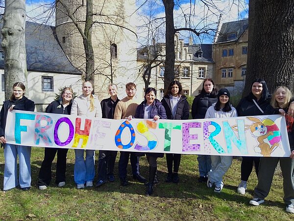 Eine Gruppe von Schülern steht auf einer Wiese vor schlossartigen Gebäuden und hält ein großes Banner mit bunten Buchstaben, auf dem Frohe Ostern steht.