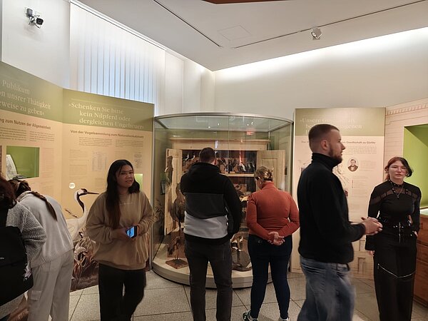 Praktische Übung zur Gesprächsführung: Ein Erfahrungsbericht aus dem Senckenberg Museum Eine Gruppe von Menschen in einem Museum, umgeben von Ausstellungstafeln und einem gläsernen Schaukasten mit ausgestopften Vögeln. Die Wände sind mit informativen Texten versehen, und einige Besucher schauen sich die Exponate interessiert an.