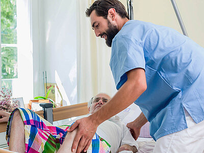 Das Bild zeigt einen hellen Raum mit einem großen Fenster. Eine Person liegt im Bett, bedeckt mit einer bunten gestreiften Decke. Ein Pfleger in blauer Uniform assistiert der Person. Grüne Pflanzen sind auf dem Fensterbrett zu sehen.