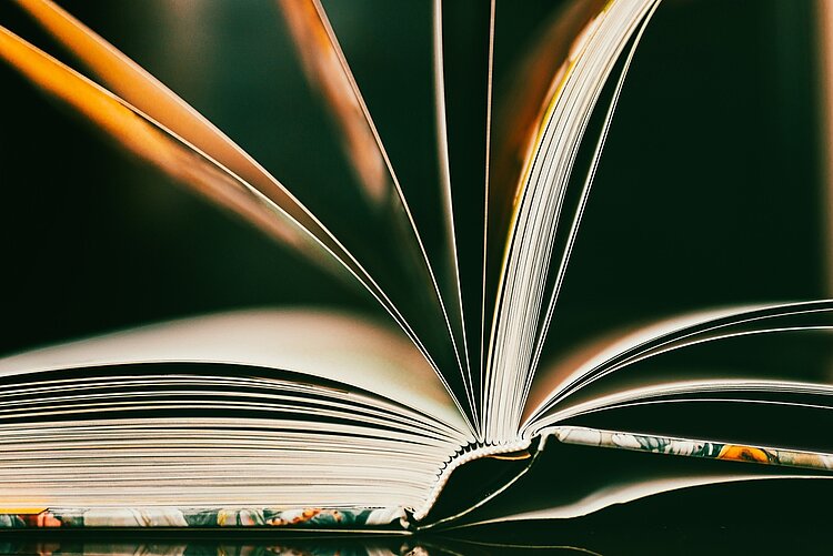 Ein offenes Buch mit leicht gewellten, weiß-gelben Seiten, das im Vordergrund liegt. Die Seiten sind leicht aufgefächert, sodass ein dynamisches Muster aus Linien und Schatten entsteht. Der Hintergrund ist dunkel und unscharf, was den Fokus auf die Struktur und Textur der Buchseiten lenkt.