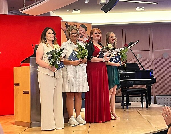 Feierlicher Abschied: Exmatrikulation der Absolvent*innen 2025 Vier Frauen stehen auf einer Bühne und halten Blumen und Zertifikate. Sie sind elegant gekleidet und lächeln. Im Hintergrund ist ein rotes Banner und ein Klavier zu sehen.