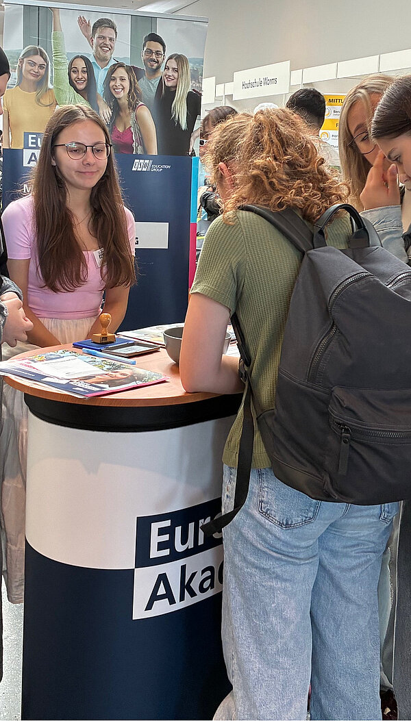 Euro Akademie Mainz auf BIM Rheinhessen Eine Schülerin der Euro Akademie Mainz berät eine Messebesucherin an einem Informationsstand. Im Hintergrund sind weitere Interessent*innen zu sehen, die sich über die Angebote der Akademie informieren. Der Stand ist mit dem Logo der Euro Akademie und Informationsmaterial ausgestattet.