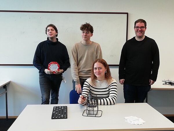 Vier lächelnde Schüler in einem Klassenraum: drei junge Männer stehen hinter einer jungen Frau an einem Tisch, auf dem eine kleine Lostrommel steht.