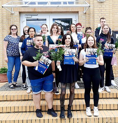 Für das Gruppenfoto haben sich die Absolvent*innen vor das Schulhaus der Euro Akademie Bitterfeld-Wolfen auf den Stufen platziert. Sie halten stolz ihre blauen Mappen mit den Zeugnissen in die Kamera. Außerdem hat jede der ehemaligen Schüler*innen eine rosafarbene Gerbera in der Hand.