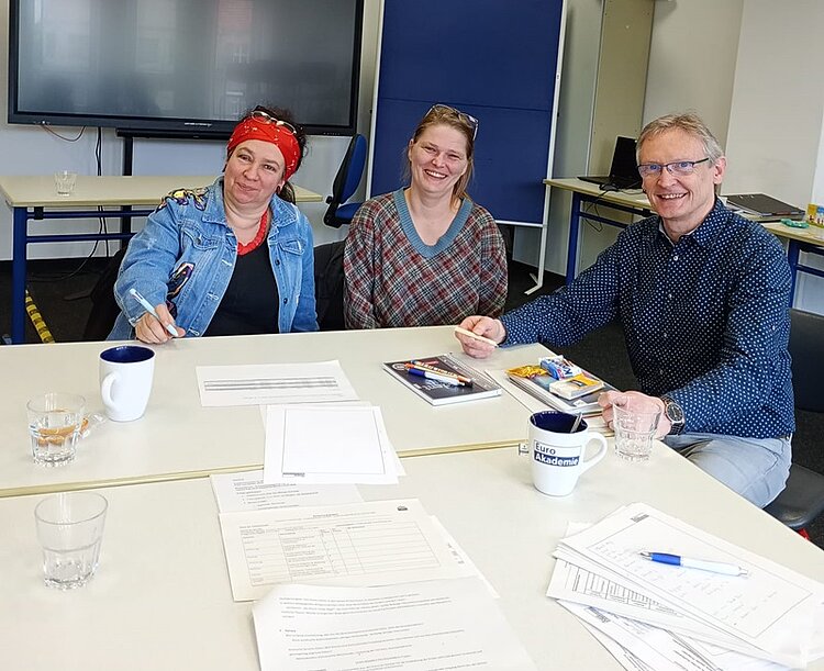 Drei Personen sitzen an einem rechteckigen Tisch in einem Besprechungsraum. Auf dem Tisch liegen mehrere Blätter, ein Notizbuch, ein Kugelschreiber, eine Packung Kekse, drei Tassen und zwei Gläser mit Wasser. Im Hintergrund steht ein großer Monitor auf einem Rollwagen sowie ein weißes Regal. Die Personen tragen Freizeitkleidung und blicken in Richtung Kamera.