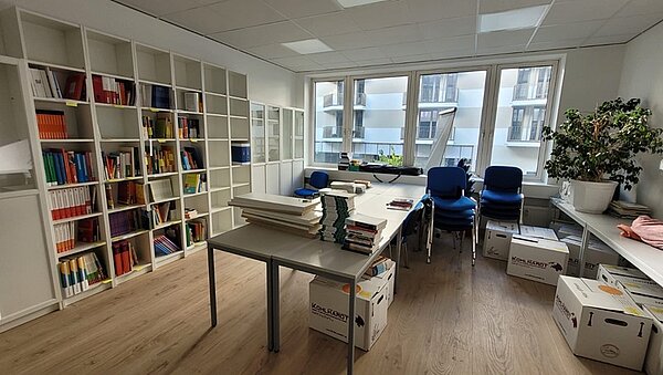 Blick in einen großen, hellen Raum. Links eine Bücherregalwand in weiß, mittig stehen zusammengeschobene Tische, an der Fensterfront stehen gestapelte Polsterstühle mit blauen Bezügen, rechts weitere weiße Tische, auf denen eine große Grünpflanze steht. Auf dem Boden direkt darunter stehen mehrere Kartons.