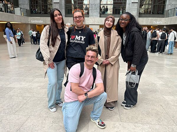 Fünf lächelnde Personen posieren gemeinsam für ein Foto in einer hellen, öffentlichen Halle. Im Hintergrund sind Schilder mit den Aufschriften ‚Garderobe‘ und ‚Tickets‘ zu sehen, was darauf hindeutet, dass sie sich in einem Kultur- oder Veranstaltungsort befinden.