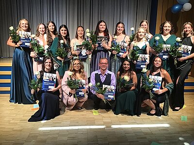 Fünfzehn junge Frauen in festlichen Kleidern und ein ebenfalls festlich gekleideter junger Mann in einer Weste stehen bzw. knien auf einem Holzparkett vor einer Bühne. Sie haben Zeugnismappen und weiße Rosen in der Hand. Die Bühne im Hintergrund ist mit blauen und weißen Lufballons geschmückt.