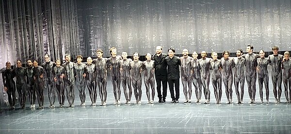 Eine Gruppe von Balletttänzerinnen und -tänzern steht in einer Reihe auf der Bühne des Opernhauses Hannover. Sie tragen eng anliegende, silbergraue Kostüme mit einem metallischen Schimmer, die ihre Körperformen betonen. Die Bühne ist mit einem Vorhang aus silbrig glänzendem Stoff im Hintergrund ausgestattet, der eine elegante Atmosphäre schafft. In der Mitte der Reihe stehen zwei Personen in schwarzer Kleidung, die sich von den Tänzern abheben. Die Tänzerinnen und Tänzer halten sich an den Händen und blicken nach vorne. Die Szene zeigt den Schlussapplaus des Balletts „Goldberg“, das von Schülerinnen und Schülern der Euro Akademie Hannover besucht wurde.