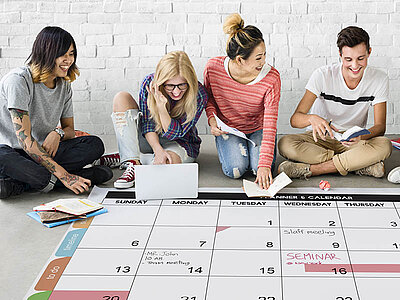 Vier Personen sitzen auf dem Boden mit einem geöffneten Laptop und einem übergroßen Kalender und planen ihren Zeitplan.