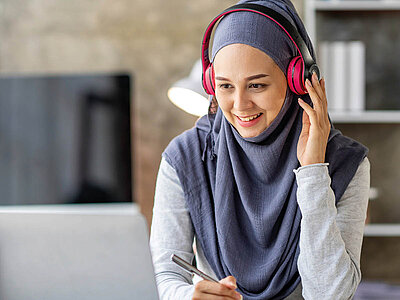 Eine lächelnde Frau mit Kopfhörern und einem grauen Hijab sitzt an einem Schreibtisch und hält einen Stift in der Hand. Im Hintergrund sind ein Laptop und Bücherregale zu sehen.