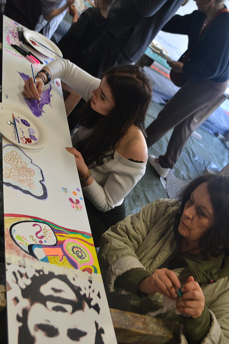 Ein Tag voller Farben und Geschichten Mehrere Personen malen konzentriert und kreativ gemeinsam auf einem langen, schmalen Streifen Papier. Im Vordergrund sitzt eine jüngere Person, die mit intensiver Konzentration ein lilafarbenes Motiv malt. Neben ihr sitzt eine ältere Person, die ebenfalls malt. Das Bild enthält bereits farbenfrohe und abstrakte Muster, Symbole und Formen.
