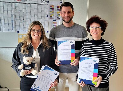Drei Personen posieren mit ihren Zertifikaten. Die Person in der Mitte ist ein Mann in einem grauen Pullover. Links von ihm steht eine Frau mit langen blonden Haaren und einer Brille., die ein Zertifikat hält und einen Teddybären mit einem "Euro Akademie" T-Shirt in der Hand hat. Rechts steht eine weitere Frau mit kurzen braun-roten Haaren, einer Brille und einer schwarz-weiß gestreiften Pullover. Im Hintergrund ist ein Kalender für das Jahr 2025 zu sehen.