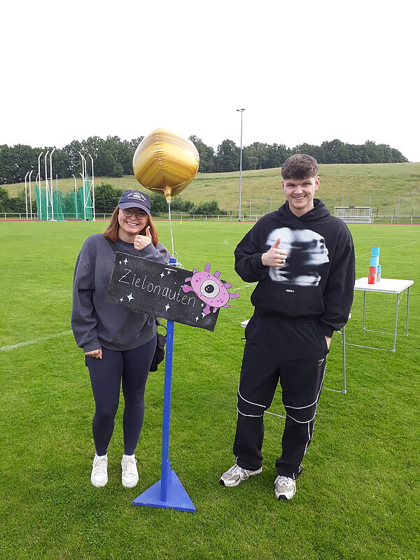 Unser Motto zum Sportfest: "Auf zu neuen Galaxien!" Zwei junge Erwachsene stehen auf einem Sportplatz neben einem Schild mit der Aufschrift „Zielonauten“. Sie lächeln und zeigen den Daumen nach oben. Ein goldener Ballon ist an das Schild gebunden.
