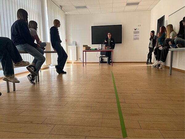 SCHAU REIN! – Woche der offenen Unternehmen Sachsen Ein heller Seminarraum mit jungen Menschen, die an den Seiten auf Tischen sitzen, während vorne eine Person steht und spricht. Auf dem Boden ist ein grüner Streifen als Markierung.