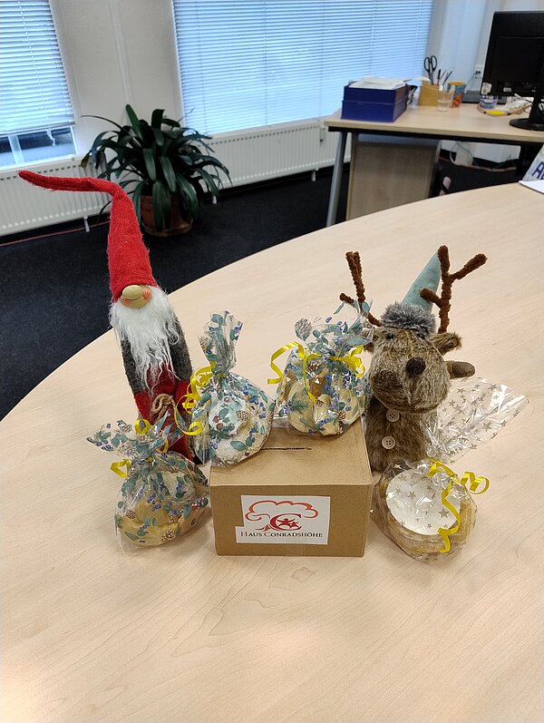 Auf einem Schreibtisch stehen ein Weihnachtswichtel und ein Plüschrentier neben einer Spendenbox „Haus Conradshöhe“, umgeben von mehreren durchsichtigen Geschenkbeuteln mit gelben Schleifen.