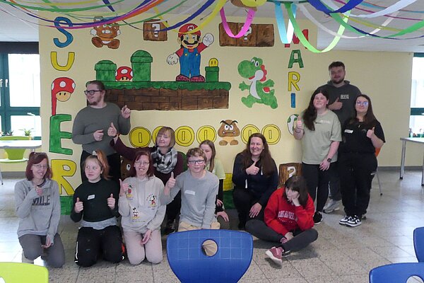 Erzieher*innen begleiten die Kreativtage an der Förderschule für geistige Entwicklung in Skäßchen Eine Gruppe von Schüler*innen und Lehrkräften posiert stolz vor der fertigen Super-Mario-Wandmalerei. Sie lachen und zeigen Daumen hoch. Bunte Luftschlangen hängen von der Decke.
