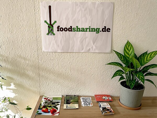 An einer hellen Wand hängt ein weißes Plakat mit dem Schriftzug 'foodsharing.de' in grüner und brauner Schrift. Links im Logo ist ein stilisiertes Besteckmotiv integriert. Darunter steht auf einer Holzoberfläche eine grüne Zimmerpflanze in einem grauen Topf. Daneben liegen mehrere Broschüren, Flyer und Karten ordentlich ausgebreitet.  Es wurde Text erkannt (OCR). Im Ergebnis wurden Eigennamen genannt. Bitte prüfen Sie das Ergebnis.  Sie erreichen bald das Nutzungslimit. Wechseln Sie in den Standard-Modus oder nutzen Sie bei hohem Bedarf das Angebot zur Stapelverarbeitung.