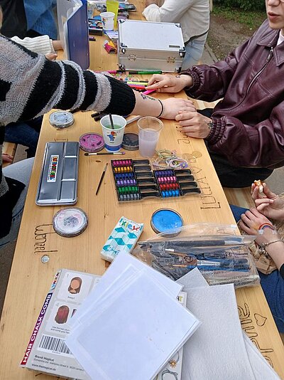 Mehrere Personen sitzen an einem Holztisch und gestalten gemeinsam kreative Bastelprojekte. Auf dem Tisch liegen bunte Stifte, Pinsel, Farbpalette, Glitzer, Aufkleber, Schablonen, Papier und andere Bastelmaterialien. Im Hintergrund sind weitere Materialien wie Haargummis und ein Behälter mit Stiften zu sehen.