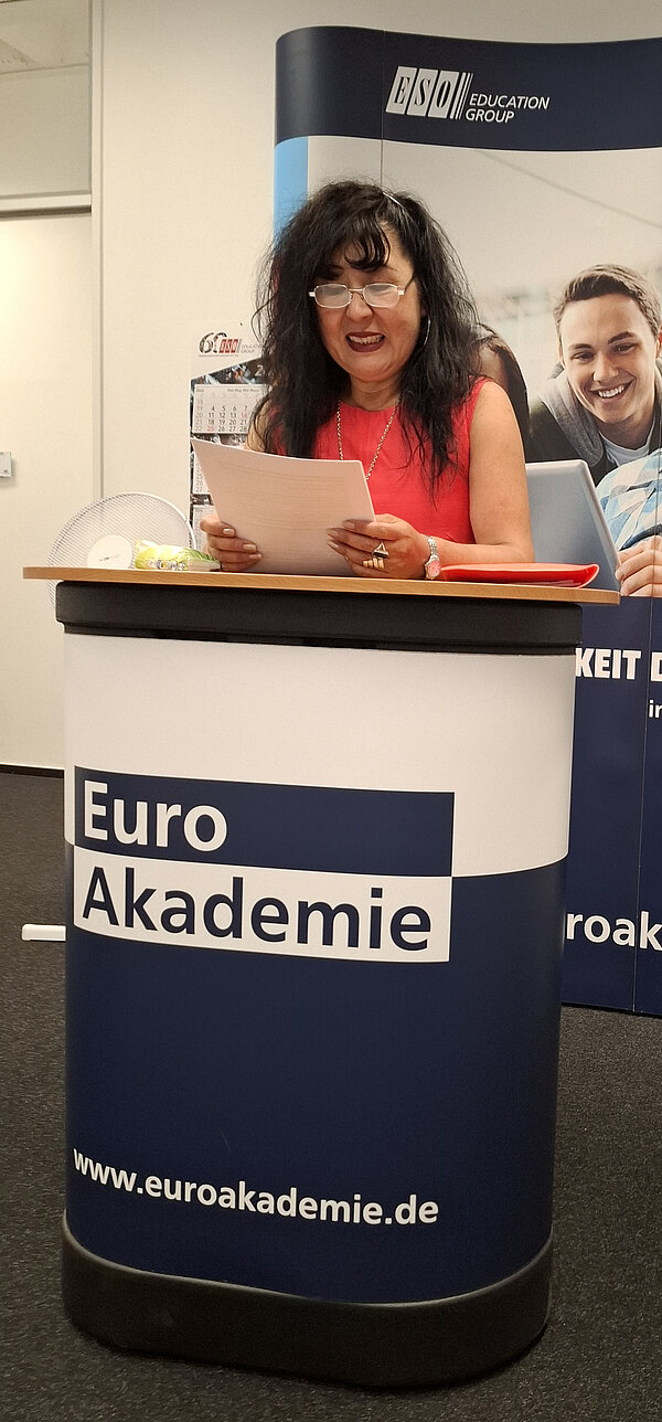 Eine Frau mit dunklem Haar und Brille steht hinter einem Podium mit der Aufschrift ‚Euro Akademie‘ und der Website www.euroakademie.de. Sie hält ein Blatt Papier in der Hand und lächelt in die Kamera. Im Hintergrund ist ein Roll-up mit dem Logo der ESO Education Group sichtbar.