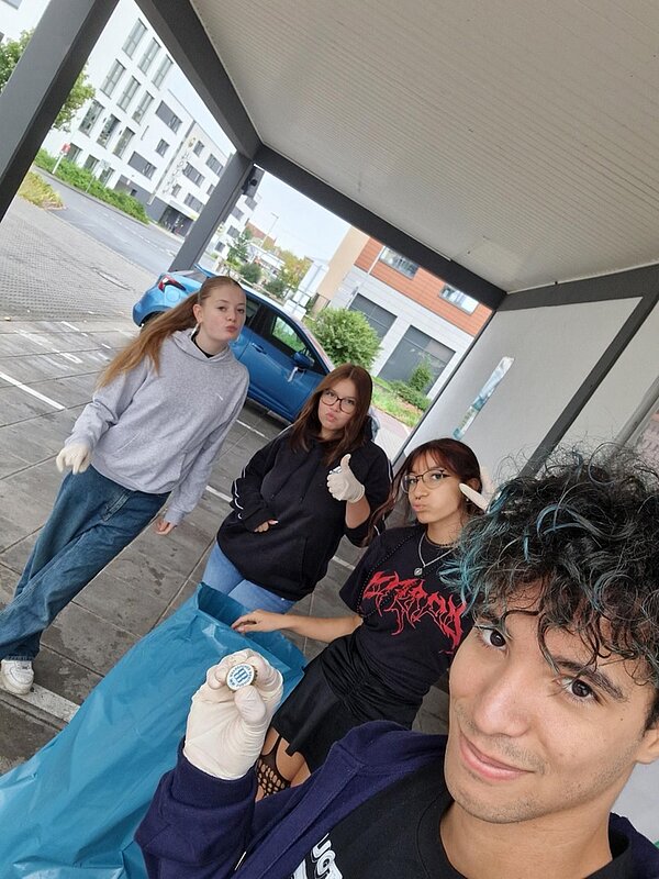Tag der sauberen Umwelt Ein Schüler hält einen Kronkorken direkt in die Kamera, dahinter stehen drei Schülerinnen mit einem blauen Müllsack. Alle Personen tragen Plastikhandschuhe.