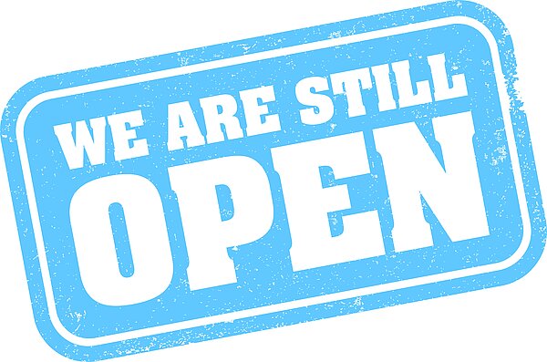 Wir haben auch in den Ferien geöffnet Auf weißem Hintergrund befindet sich ein hellblaues Schild mit dem Schriftzug "WE ARE STILL OPEN".