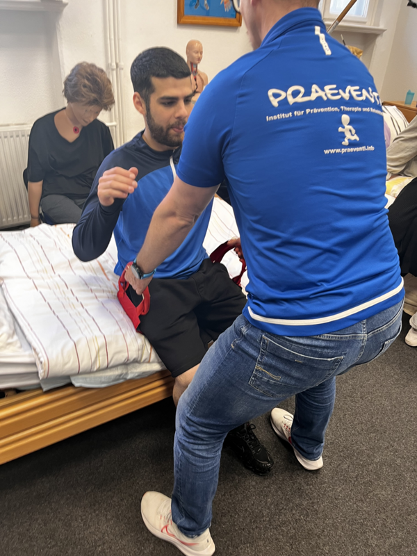 Ein Auszubildender in einem blauen Polohemd mit weißem Aufdruck unterstützt einen sitzenden Azubi beim Aufstehen von einem Bett. Die sitzende Person trägt ein dunkles T-Shirt und kurze Hosen. Im Hintergrund sitzt eine weitere Person auf dem Bett und beobachtet die Szene. Der Raum wirkt wie ein Schulungs- oder Therapieraum, da ein anatomisches Modell auf einem Schrank steht.